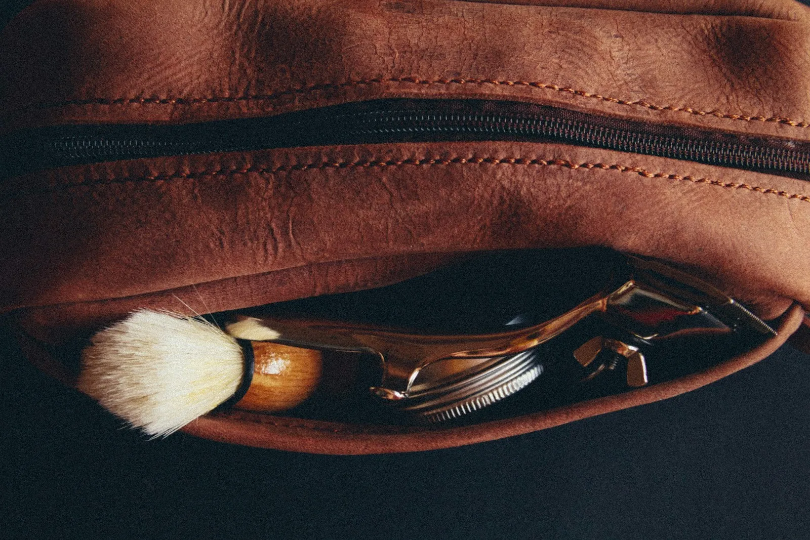 A brown leather toiletry bag lying open with a shaving brush and razor tucked into its front pocket