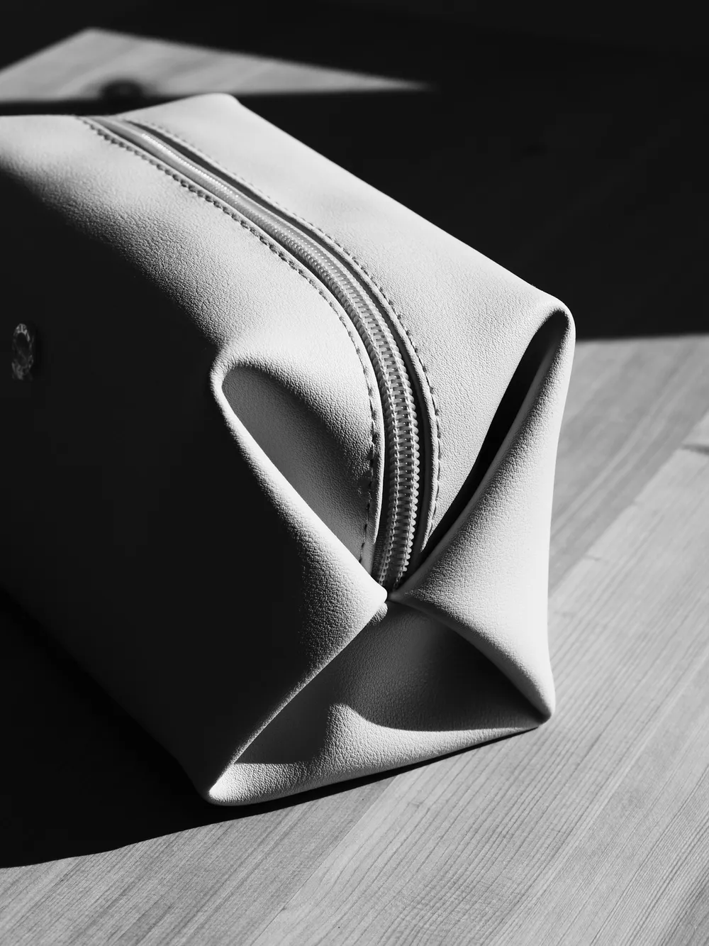 A light-colored zippered toiletry pouch sitting on a wooden table in hard sunlight